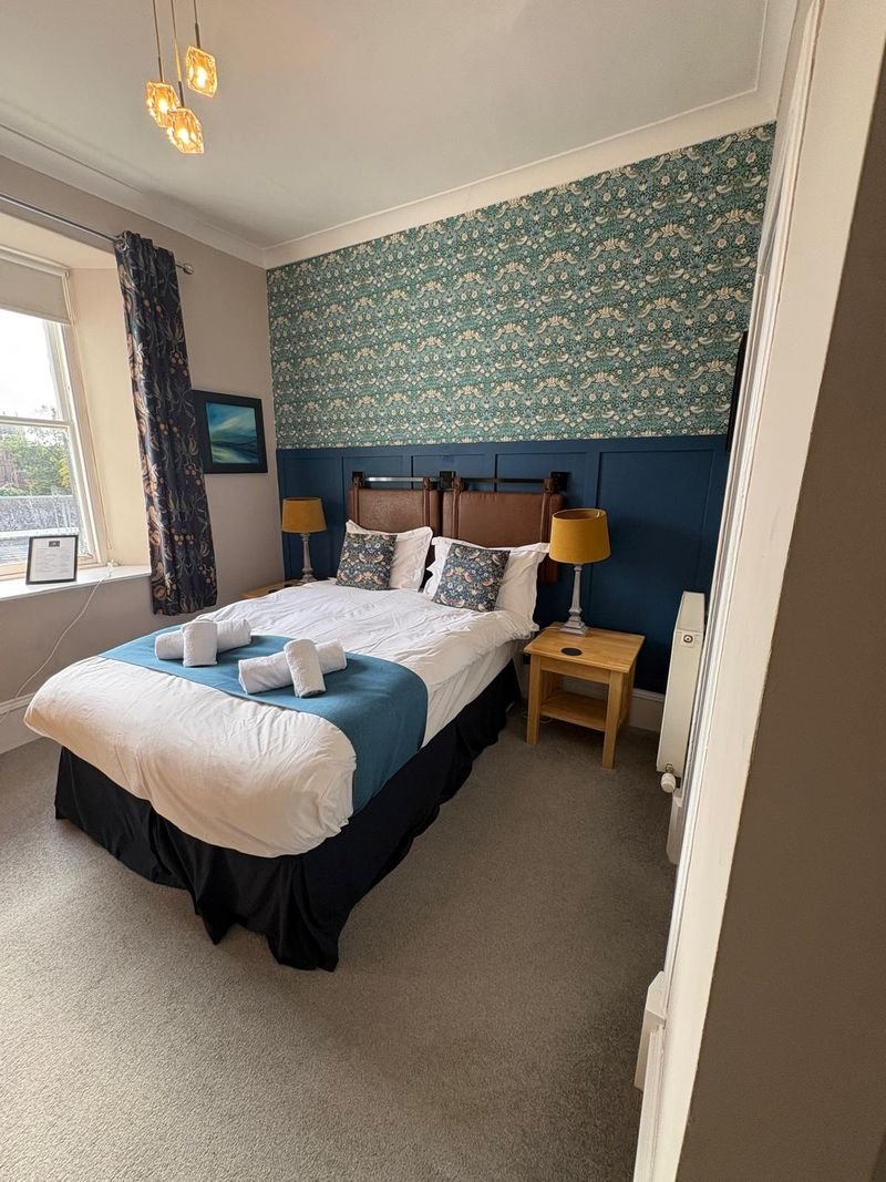 A double room with bird patterned wallpaper above navy panelling, mustard lamps and a window